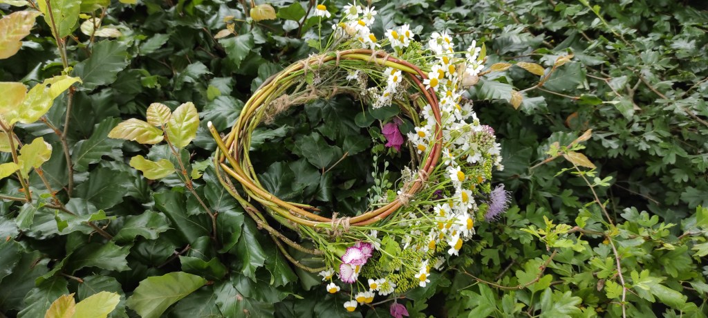 A rustic floral wreath made of various flowers and greenery, resting on lush green foliage.