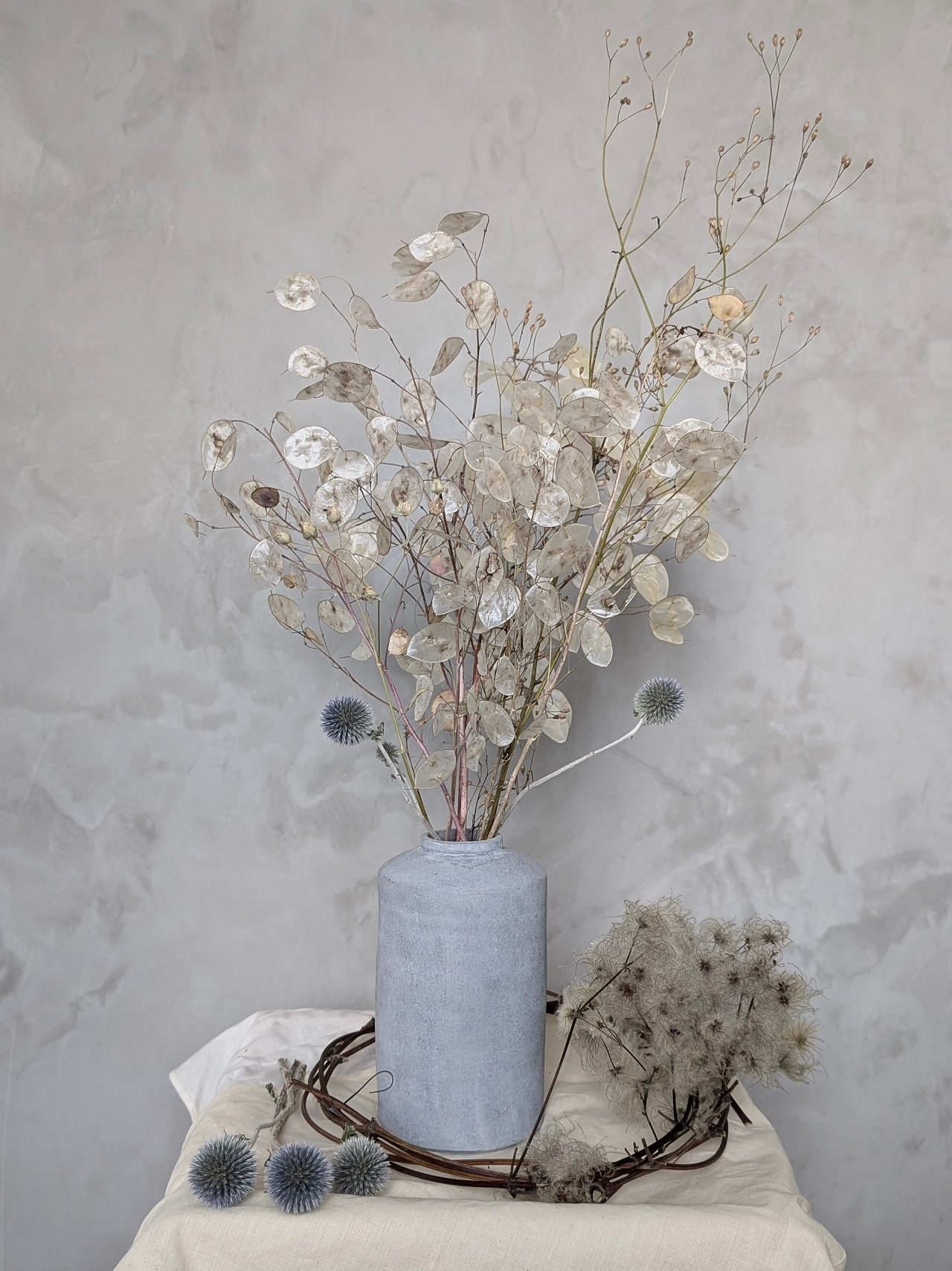 A decorative arrangement of dried flowers and foliage in a light blue vase, and lunaria, set against a muted gray background.