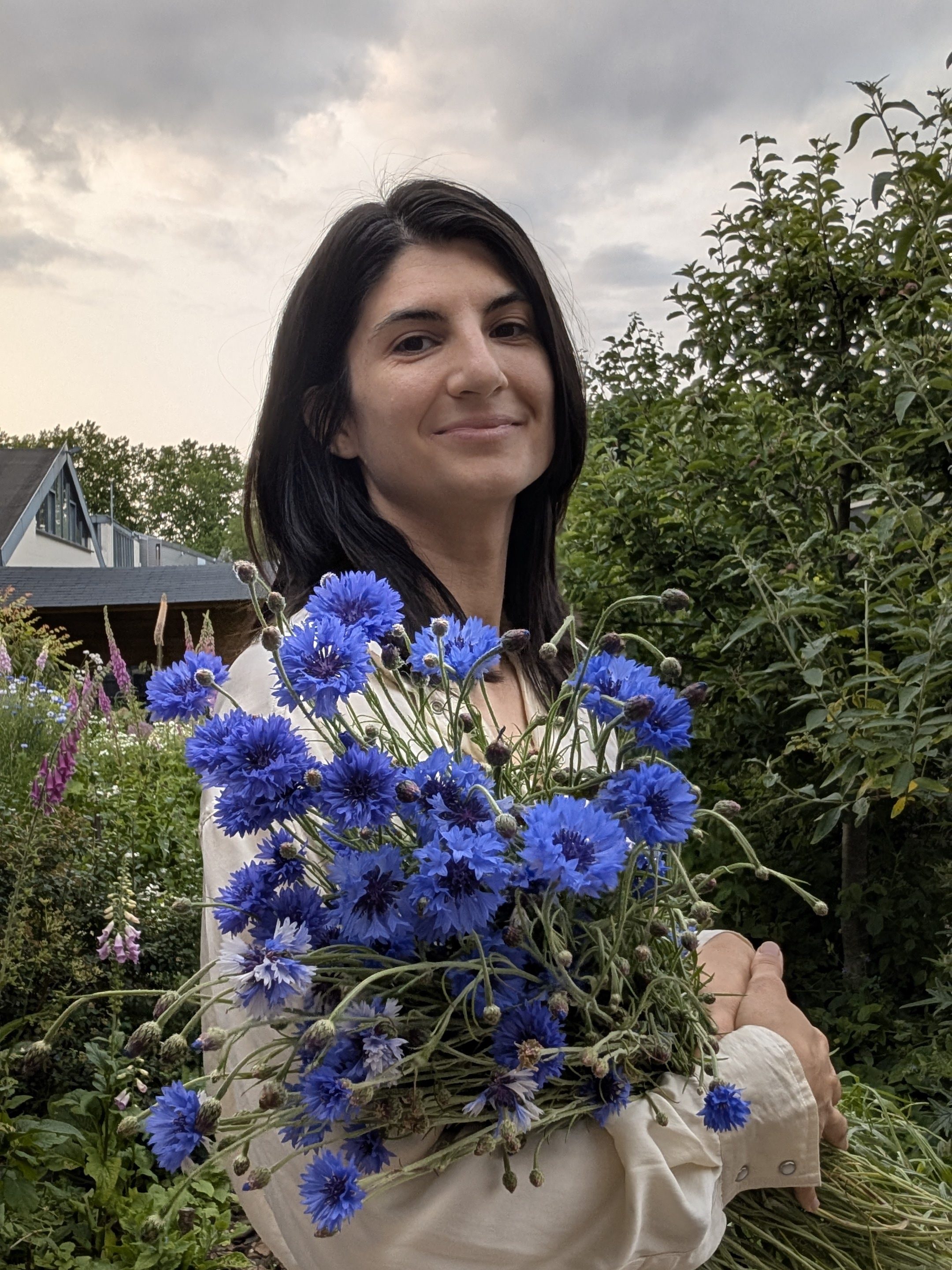 A woman smiling while holding a large bouquet of blue cornflowers in a garden setting with greenery and a cloudy sky in the background.