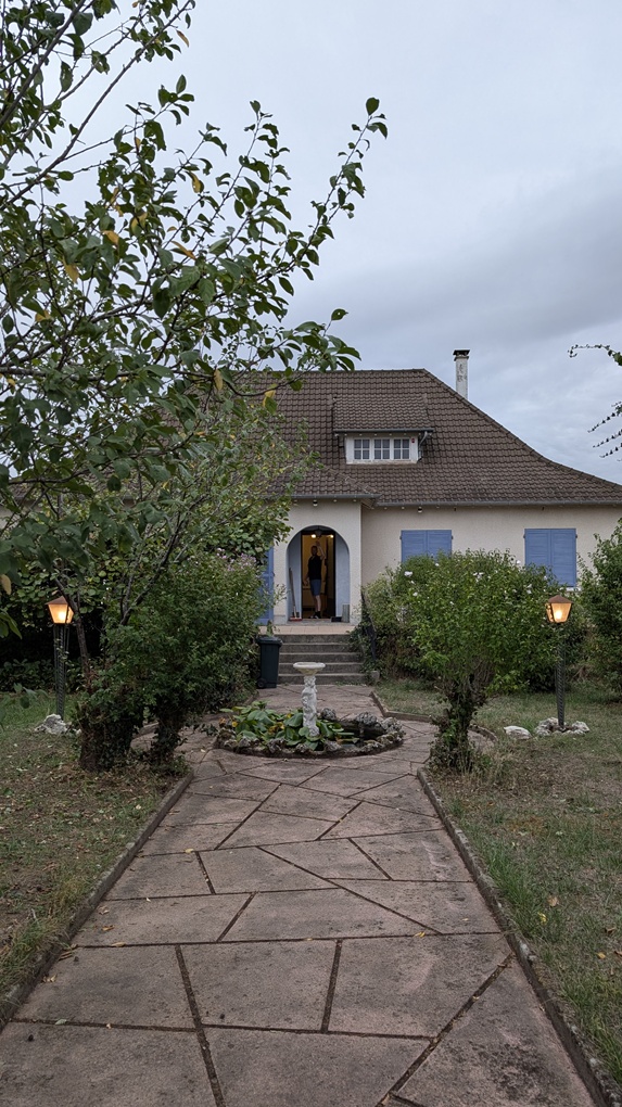 A pathway leading to a house with blue shutters and a person standing at the entrance, surrounded by greenery and garden lights.