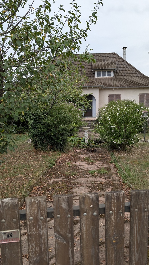 A view of a residential house with a brown roof, front door, and overgrown garden, framed by trees and shrubs, seen through a wooden gate.